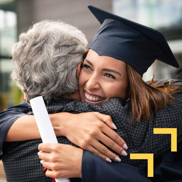 A young woman wearing a graduation hat and gown, hugging her father and smiling while holding a diploma