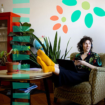 A person wearing orange socks, sitting on a chair with their legs on the coffee table, looking at a distance, smiling.